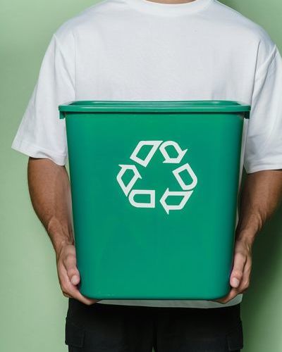 Auf dem Bild ist der Oberkörper einer männlichen Person mit weißem T-Shirt und schwarzer Hose zu sehen, die eine grüne Box mit dem Recycling-Symbol in den Händen hält.